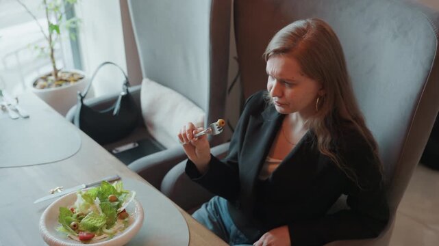 White woman reacting to salad taste, creative consultant tries new dressing and grimaces, pushes fork away while seated by window with handbag and plant, casual jacket, closeup facial expression