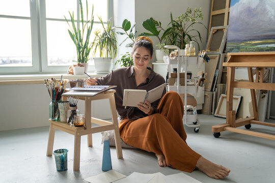 Happy woman creator enjoying art hobby and drawing pictures in notebook using watercolors mediums, cheerful female looking through sketchbook smiling, finding inspiration for new creative projects.