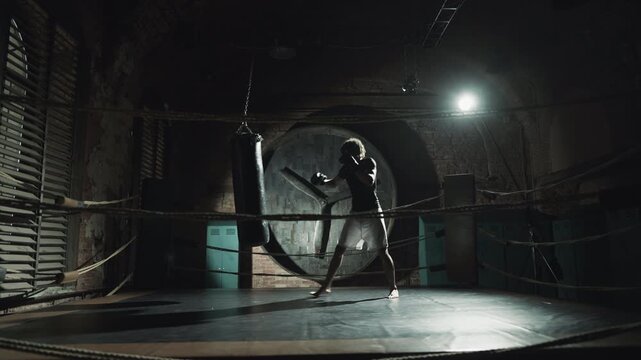 African american athlete practicing box in dark gym, hitting punching bag. Kickboxing and combat sports, self-defense class for men, professional sportsman training alone in night, future champion
