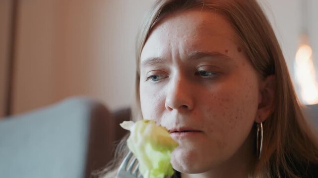 Caucasian woman grimacing after bite closeup reaction shot of caucasian woman chewing salad with bitter expression, fork near mouth, freckled skin, tense jaw, indoor couch setting, candid raw taste