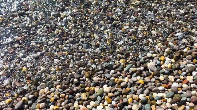 Varied colorful pebbles form a wet, textured shoreline as gentle seawater washes over stones, creating a tranquil, full-frame coastal pattern in bright daylight