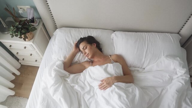 Young woman sleeping peacefully in a soft white bed, calm bedroom filled with gentle morning light. Concept deep rest and home comfort.
