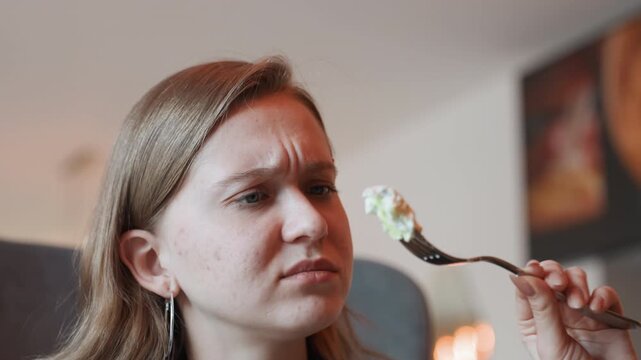 Caucasian woman grimacing at forkful salad, closeup reaction in intimate bistro setting, inspecting greens and dressing with visible freckles and concern, reluctant taste test under soft ambient light