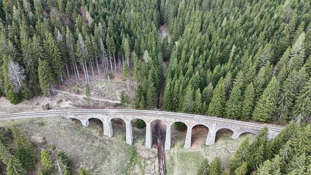 Aerial view tracking along the historic Chmarossky viaduct. This arched stone railway bridge stands among a dense pine forest in Telgart, Slovakia