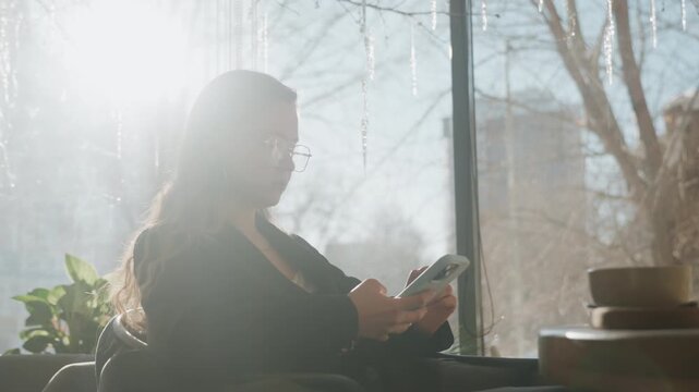 White woman silhouetted by winter window checking smartphone, contemplative strategist mood, soft backlight and city skyline beyond glass, quiet cafe scene, focused planning and reflective atmosphere.