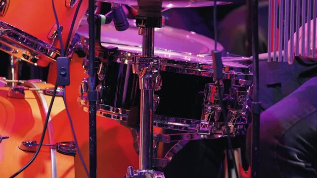 A close-up of a professional drum kit during a live jazz concert. The stage is illuminated by bright purple and orange stage lights. The drummer plays with drum brushes