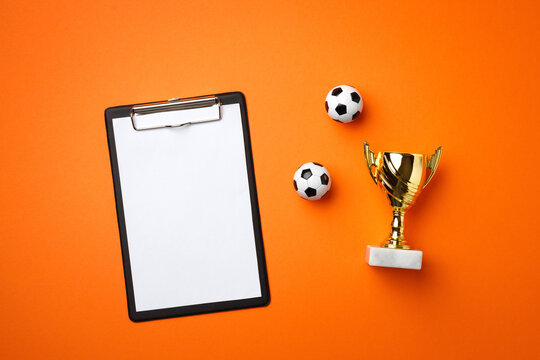 Blank clipboard, golden trophy and two mini balls on bright orange background create a winner&rsquo;s victory championship setup composition. Top view, flat lay.