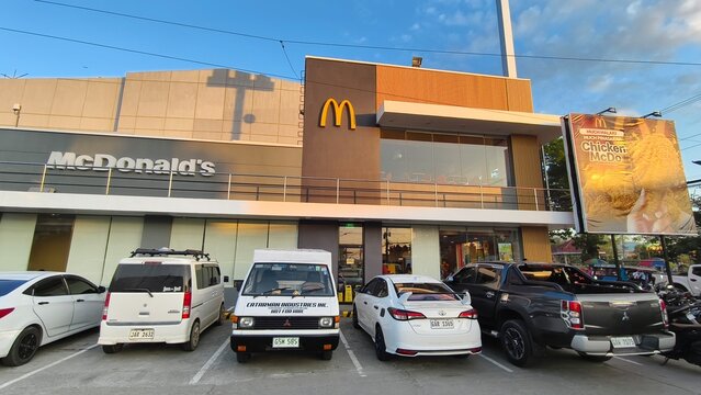 Tubigon, Bohol, Philippines - McDonald's modern branch exterior with parked cars and large fried chicken billboard at sunset.