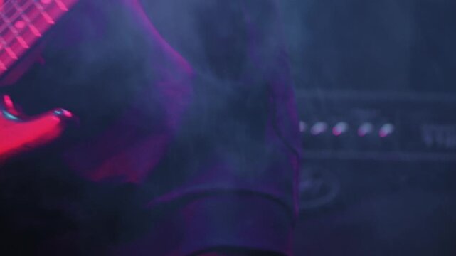 Close-up of a bassist's hands on a bass guitar during a live performance. The energetic scene is lit with dramatic pink and blue stage lights, creating a moody rock and roll atmosphere