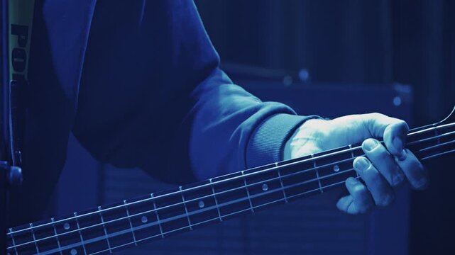 Close-up of a bassist's hands on the fretboard of a bass guitar. Moody, low-key lighting highlights the musician's fingers. Perfect for music, concert, and band concepts