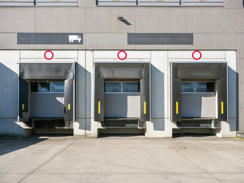 Three loading dock bays with grey doors and red numbered signs at a modern logistics warehouse goods entry.