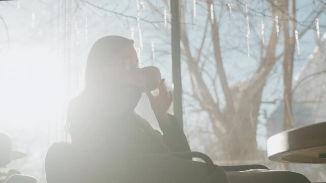 White woman silhouetted against bright window, strong backlight and gentle lens flare, winter trees visible outside, contemplative sip while seated in armchair, cinematic contrast and serene