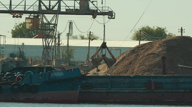 Industrial scene at river port. Large gantry crane with clamshell bucket loads sand onto cargo barge. Gritty, realistic view of shipping, logistics, and bulk material transport on sunny day