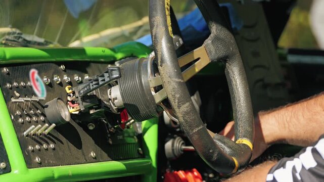 Close up view of racer hands on steering wheel inside custom built all-terrain vehicle. Dusty dashboard with toggle switches and green roll cage. Focus on action and preparation for competition