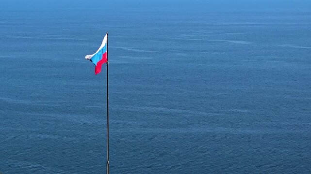 Russian flag on a tall flagpole rippling in the breeze above calm blue sea, serene coastal horizon stretching into the distance under clear daylight sky, peaceful and patriotic