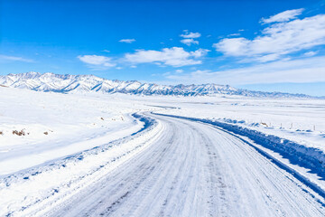 Obraz premium A winding snow-plowed road cuts through a vast frozen snowy plain, leading towards snow-capped mountain ranges under a bright blue cloudy sky.
