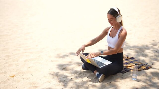 woman in activewear using computer while sitting on tropical beach, work from anywhere concept.