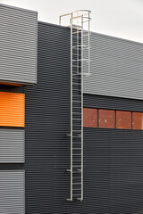 Architectural facade with orange cladding corrugated metal panels, industrial vertical ladder with a safety cage or crinoline, large glass window. Building with vertical relief and material contrast. © R_Yosha
