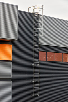Architectural facade with orange cladding corrugated metal panels, industrial vertical ladder with a safety cage or crinoline, large glass window. Building with vertical relief and material contrast.