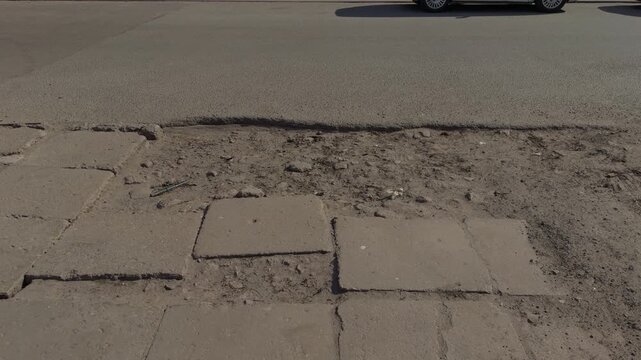 Patched road surface with passing vehicle visible at top, depressed paving squares and uneven tarmac indicate repeated repairs, urban traffic context with textured foreground detail