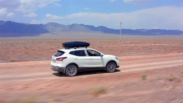 Smooth side tracking drone shot of white SUV cruising along dusty rural road in Kazakhstan steppe, distant mountain ridge, dry soil textures, sparse shrubs, open sky, cinematic travel motion