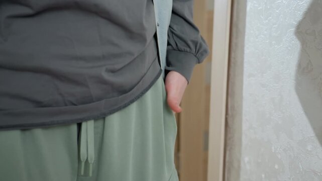 Closeup hands in pocket denim shirt, relaxed model adjusts green joggers and oversized sweater near doorway, tactile fabric and drawstring visible, soft natural light, styling moment for loungewear