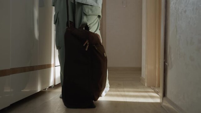 Woman entering narrow apartment hallway with suitcase, pulling wheeled bag toward closet, soft morning light casting long shadows, quiet mood, new tenant arriving, tentative steps toward open door