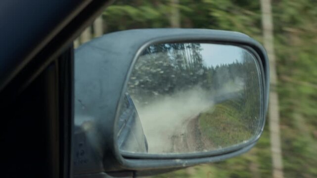 Mirror reveals dust. Side mirror captures gravel cloud ahead on wooded narrow path. Reflective surface displays trailing dust plume during urgent drive through dense wilderness