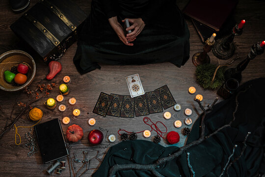 A witch fortune teller casts a spell on Tarot cards while sitting on the floor among candles and pumpkins. A sorceress on Halloween night.