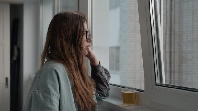 White woman on phone window sipping tea from cup on windowsill side profile gaze toward city buildings wearing denim and sweater thoughtful conversation overcast light indoor apartment calm longing
