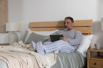 Fototapeta premium Mature man sitting on bed wearing pajama pants, holding tablet and looking right