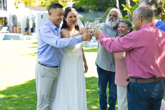 Bride in white gown and veil raising glass and initiating wedding toast, guests clinking on lawn