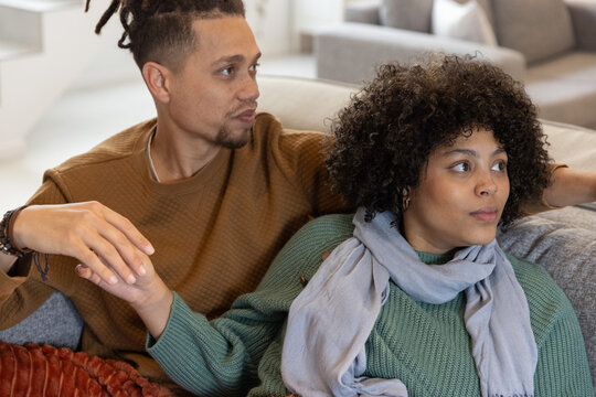 Diverse couple sitting on gray sofa in modern living room, holding hands, wearing knit sweaters