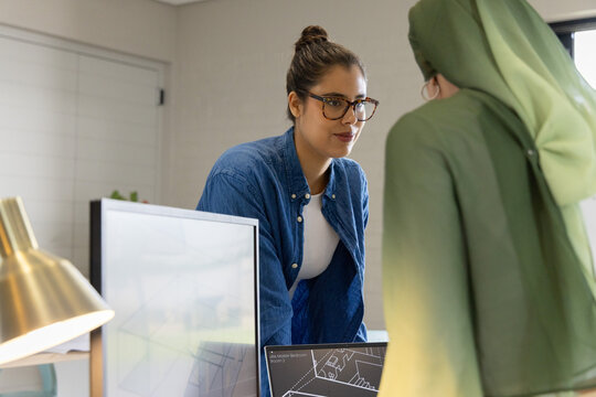 Diverse female coworkers leaning over office desk studying CAD on monitors, glasses and green hijab