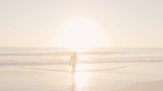 Surfer walking into surf at sunrise sunlight drawing her forward preparing board reflecting light