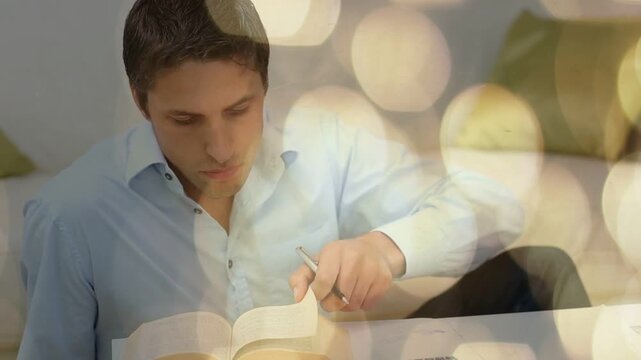 Man leaning forward, scribbling with pen, flipping pages studying open book with warm bokeh overlay