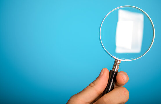 Close-up of a hand holding a magnifying glass, revealing a blurred white object against a vibrant bl