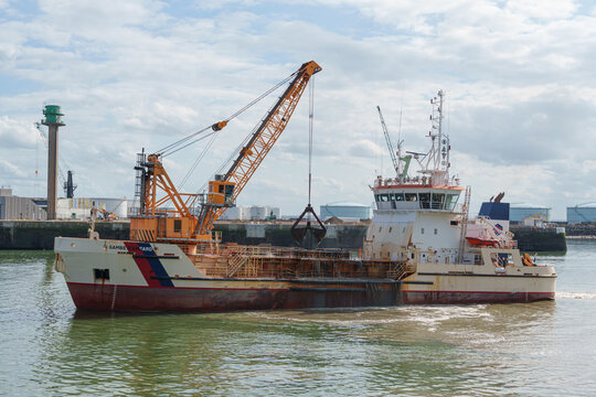 Large dredging vessel sails through an industrial harbor under cloudy skies
