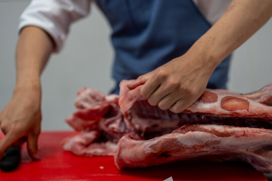 Gros plan sur un boucher en tablier bleu d&eacute;sossant avec pr&eacute;cision une &eacute;paule d'agneau sur une planche rouge, illustrant un savoir-faire artisanal et professionnel.