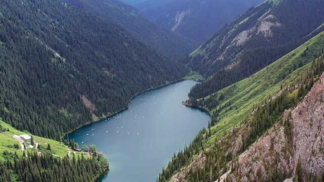 Drone flying backward over mountain lake, pulling away from Kolsai lakes in Kazakhstan with steep slopes and forested hills, expanding panorama revealing travel destination and remote nature landscape