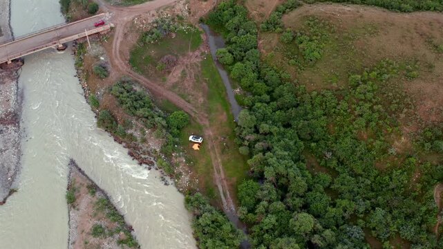 Bridge crosses flowing river as drone rises vertically revealing Chilik River landscape with road junction, greenery and hillside terrain in Kazakhstan countryside aerial view