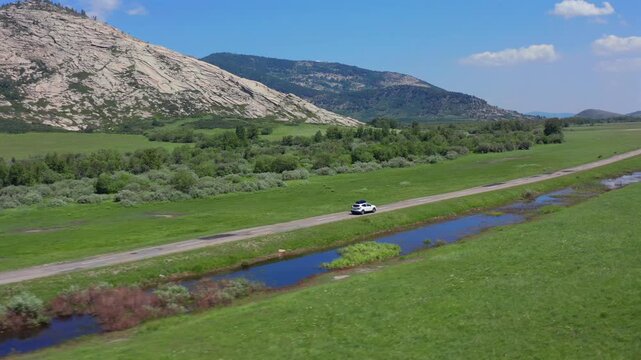 Aerial drone view of car driving along rural road through green valley with small river and rocky mountains in Kazakhstan, cinematic travel journey concept, exploration, freedom and scenic nature