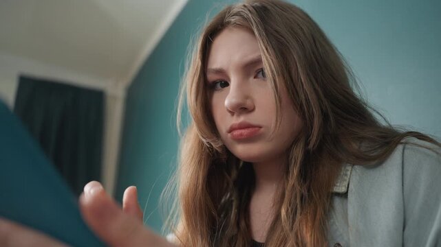 Young woman using tablet in bedroom, pensive student reviewing notes against teal wall, lowangle closeup with soft lamp glow, casual sweater, long hair, subtle shifts from focus to faint smile, quiet