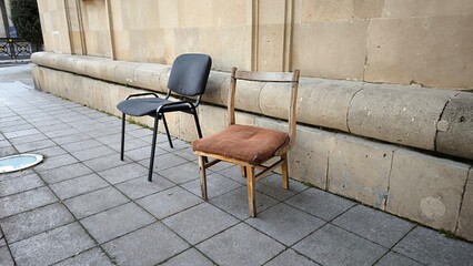 Contrast concept with vintage wooden chair and modern office chair. An old wooden chair with a brown cushion stands next to a contemporary black office chair against a stone wall. This composition hig © Amina
