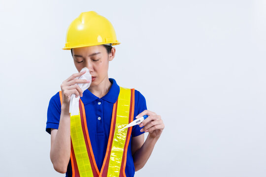 Sick Asian female engineer in yellow hard hat and safety vest, holding a thermometer and tissue, isolated on white background. Concept of fever flu or occupational health issues.