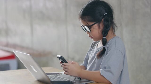 Young girl wearing glasses using a smartphone while sitting at a table with a laptop in a modern workspace environment