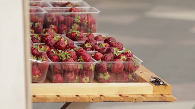 Vibrant red strawberries, still adorned with green leafy tops, are arranged in clear plastic containers. The setup sits on a rustic wooden stand, evoking a fresh market atmosphere.