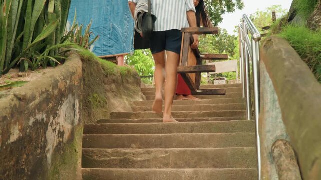 Bare feet carrying wooden chair up weathered concrete steps, closeup on legs and hands, striped shirt man ascending, mosscovered walls, tactile movement and balance in sunlit tropical setting.
