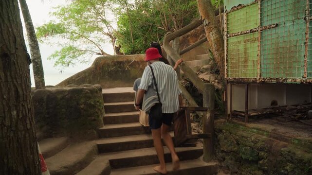 Couple climbing mossy beach stairs, playful cove discovery with man leading up stairway carrying tote, candid vlog style travel moment, rusted door and wooden railing, breeze through palm foliage,