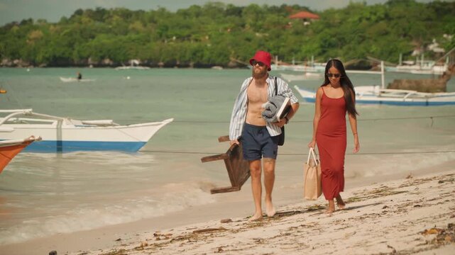 Couple walking on beach under sun, woman in red dress, man in red hat carrying wooden tea box and suitcase, outrigger boats moored offshore, green headland, gentle waves, warm cinematic tones,